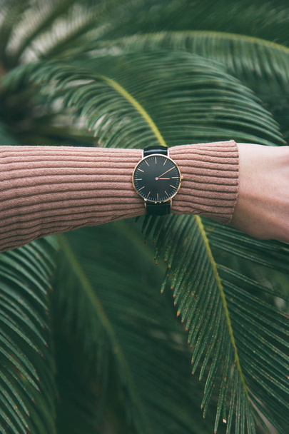 ideal fashion outfit accessories. trendy fashion blogger wearing an elegant black and golden watch. close up fashion details. palm leaf in the background. - Photo, Image