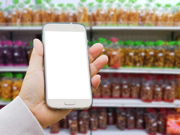 main asiatique gauche avec pull tenant téléphone intelligent sur fond flou de collation faite de produits agricoles dans des emballages à vendre sur le stand du supermarché
  - Photo, image