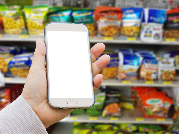 mano asiática izquierda con suéter sosteniendo pantalla en blanco teléfono inteligente sobre fondo borroso de varias marcas de frutas secas y nueces en el embalaje para la venta en el stand o estante del supermercado
  - Foto, imagen