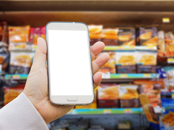 main asiatique gauche avec pull tenant écran vierge téléphone intelligent sur fond flou de diverses marques de sandwich dans l'emballage à vendre sur un stand de supermarché ou une étagère
  - Photo, image