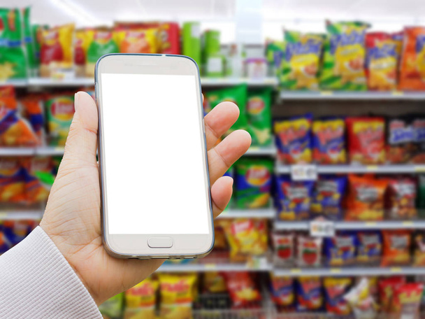 vasen aasialainen käsi villapaita tilalla tyhjä näyttö älypuhelin hämärtynyt taustalla eri tuotemerkin perunalastuja ja välipaloja pakkauksissa myytävänä supermarket seistä tai hylly
  - Valokuva, kuva