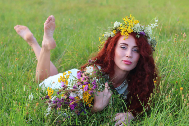 Young barefoot woman in wreath and dress lies in grass on meadow - Фото, изображение