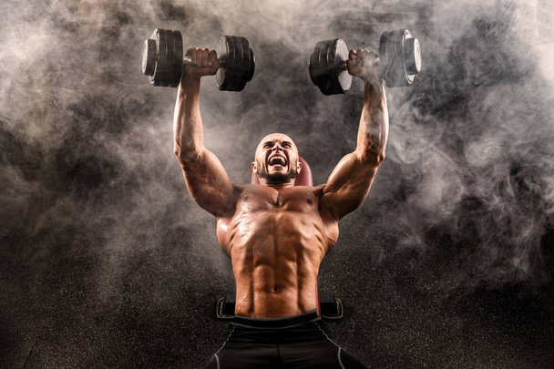 Bald Topless Muscular Man Doing Exercises With Two Dumbbells On Bench Press In Smoke - Foto, Imagen