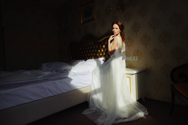 Beautiful sexy redhair lady in elegant white wedding dress. Fashion portrait of model indoors. Beauty woman with sitting on the bed. Female body. Close up bride. - Photo, Image