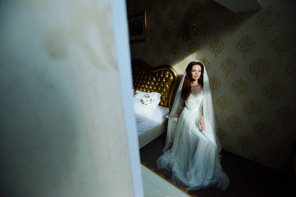 Hermosa señora pelirroja sexy en elegante vestido de novia blanco. Retrato de moda de modelo en interiores. Mujer bella sentada cerca de su cama
 - Foto, imagen