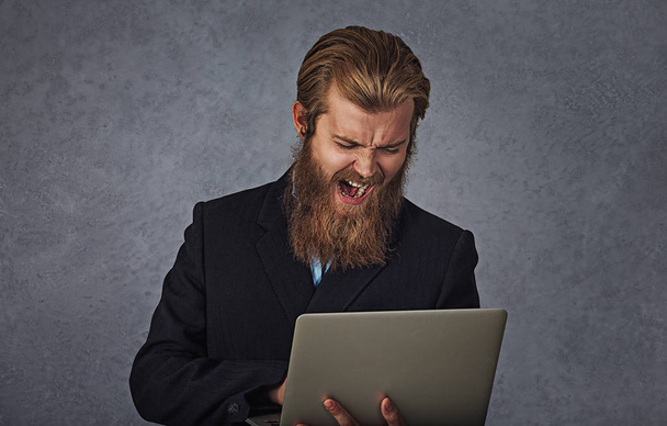 Bearded business man holding computer screaming isolated grey gray background - Foto, Bild