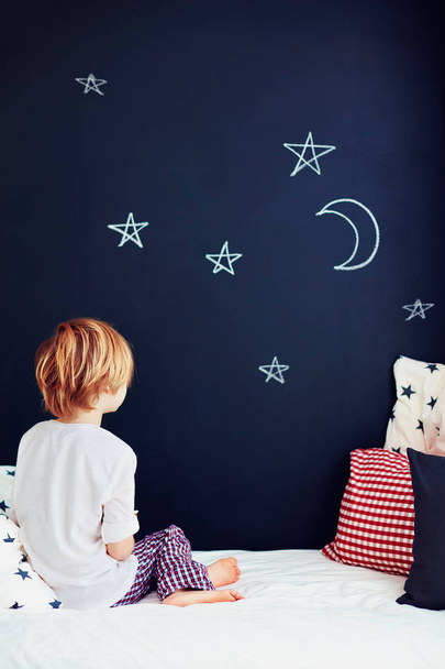 garoto bonito em pijama sonhando, enquanto sentado na cama e olhando na parede do quadro
 - Foto, Imagem