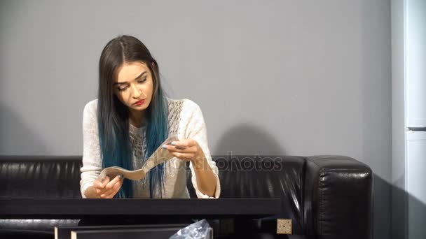 Young cute girl with colored hair sitting on a couch in their new apartment to put together a table. Shoot in kitchen. - Filmagem, Vídeo