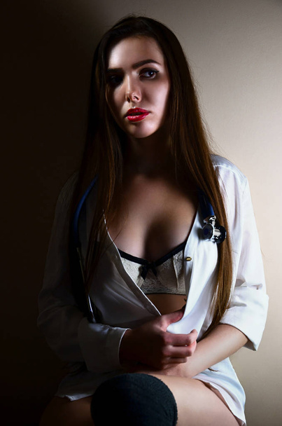 Enfermeira jovem e atraente em uma camisa branca e roupa interior branca com um estetoscópio em torno de seu pescoço sentado em uma sala com paredes bege. Estudante caucasiano sexy na escola de medicina
 - Foto, Imagem