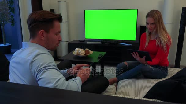 Fiatal, vonzó pár egy kanapén, egy hangulatos nappali munkát egy okostelefon és a tablet, az ember megmagyarázza valami a nő - TV-vel és egy zöld képernyő a háttérben - Felvétel, videó