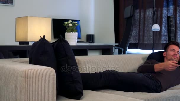 Mladý, pohledný muž leží na pohovce v útulném obývacím pokoji hodinky televize, která je mimo fotoaparát - Záběry, video