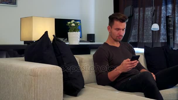 A young, handsome man sits on a couch and types on a smartphone - Materiał filmowy, wideo