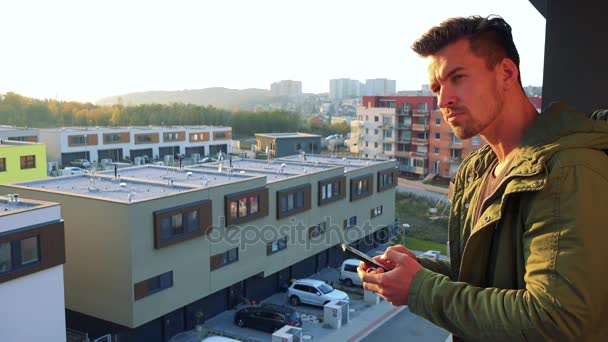 Uiteindelijk een jonge, knappe man typt op zijn smartphone op een balkon, kijkt uit - Video