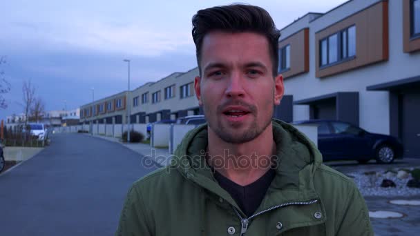 ein junger, gutaussehender Mann spricht in die Kamera über eine Straße und Gebäude hinter ihm - Filmmaterial, Video