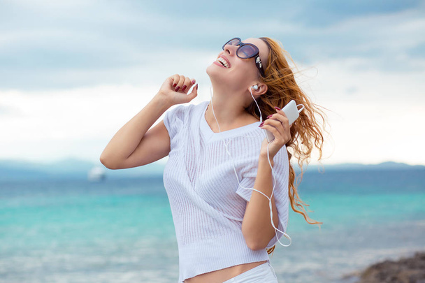 Portrait of a smiling young girl with earphones listening to music outdoors by the beach with ocean blue sky background  - Фото, изображение
