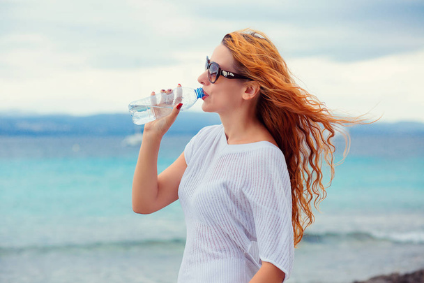 красива молода жінка в сонцезахисних окулярах п'є воду в літньому пляжі на відкритому повітрі з морським фоном
  - Фото, зображення