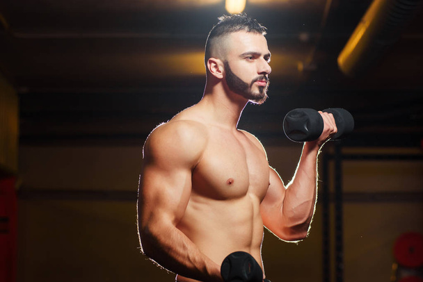 Muscular shirtless young man working out with dumbbells in gym - Foto, Bild