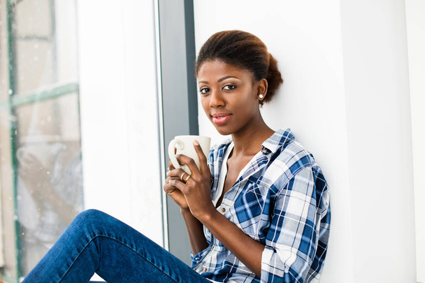 young black woman next to a window with a cup of coffee in hands - Фото, изображение