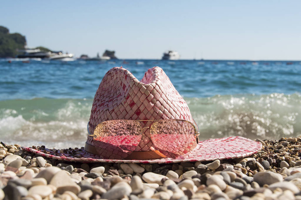 Женская соломенная пляжная шляпа с солнцезащитными очками лежит на галечном пляже, окруженном круглыми камнями на фоне синего моря с островом, волнами, лодками и яхтами. Идея для летнего отдыха на пляже
 - Фото, изображение