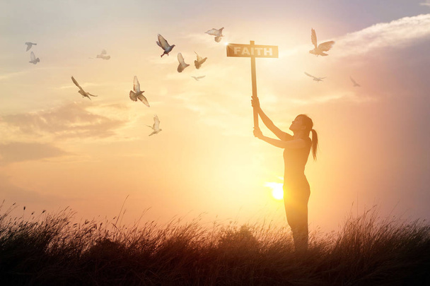 Mulher segura um sinal com palavra fé e pássaros voadores no pôr do sol da natureza
 - Foto, Imagem