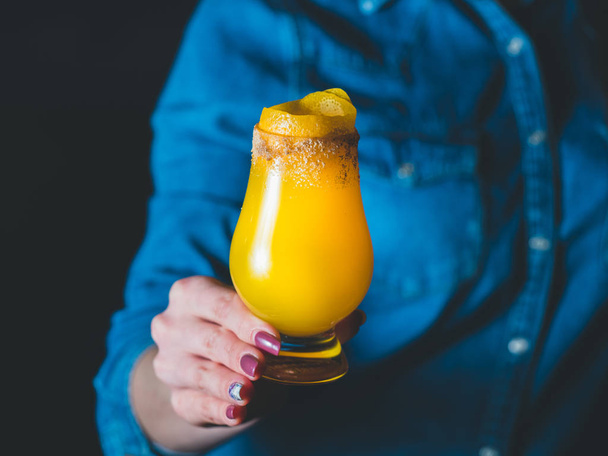 Coquetel de manga amarela com raspas de limão e crusta de açúcar em um copo de tulipa, mão feminina / barman segurando um copo com o coquetel, fundo de camisa de jeans azul. Cocktail em um centro
 - Foto, Imagem