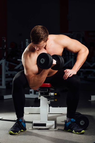 professional bodybuilder do exercises for biceps sitting on the bench, with dumbbells at gym. Big - Photo, image