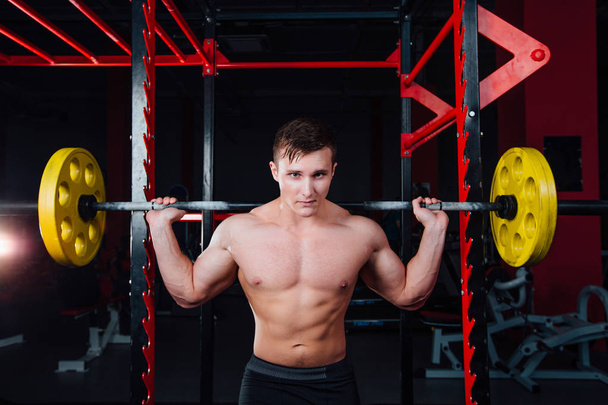 portrait of a handsome athlete at gym. man does the exercise with  barbell, standing and squatting. big confident - Фото, изображение