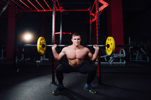 portrait of a handsome athlete at gym. man does the exercise with  barbell, standing and squatting. big confident - Фото, изображение