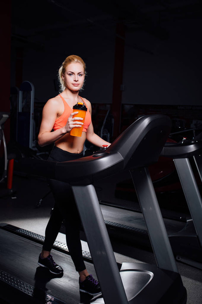 Young beautiful cute girl in fitness model in the gym running on the treadmill with bottle - 写真・画像