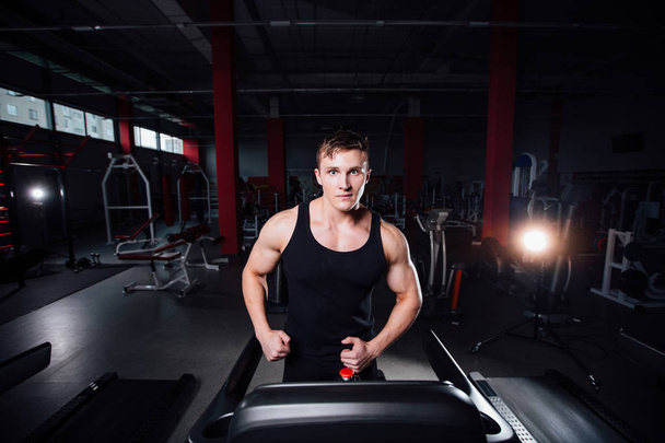 Young strong big man fitness model in the gym running on the treadmill with water bottle. - 写真・画像