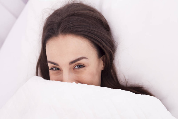 Beautiful young brunette girl waking up and smiling in a white bed. - Фото, изображение