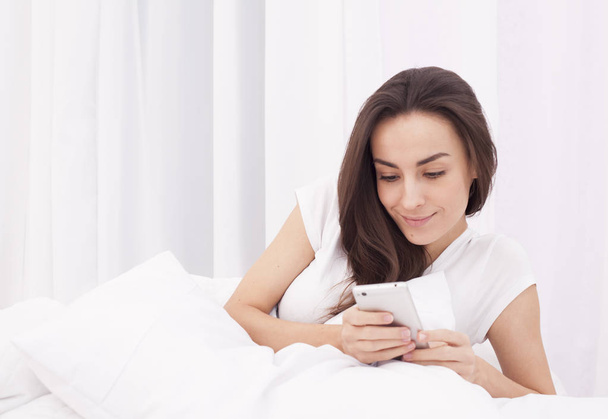Very beautiful young brunette woman writes a message on the phone in white bed. - Фото, изображение
