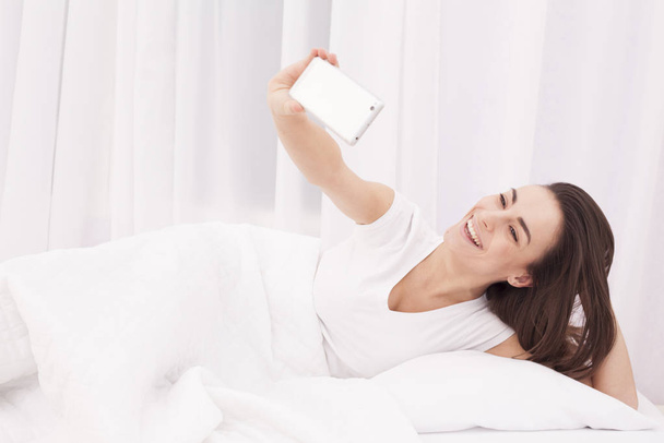 Very beautiful young brunette girl doing selfie in a white bed. - Фото, изображение