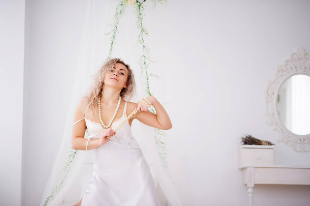 Curly blonde girl in a white silk gown boudoir robe on bed. - 写真・画像
