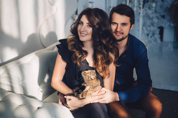 beautiful couple in love with a cat in their arms sitting on the sofa, hugging, smiling and kissing. Family concept - Fotografie, Obrázek