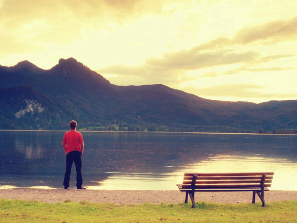Grand homme en t-shirt rouge au banc en bois sur la côte du lac des montagnes. Nuages sombres
 - Photo, image