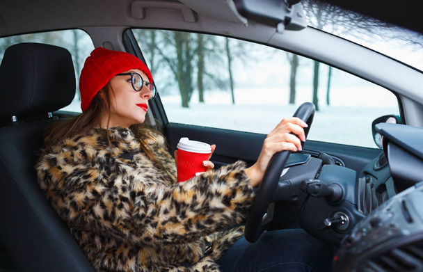 Beautiful woman in a fur coat and red hat with coffee to go driv - Фото, изображение