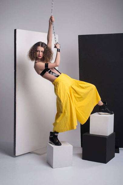 Fashion photo of beautiful girl posing in studio. Wearing yellow shorts, black boots. Straps over breast. Holding chain. Curly hairstyle. - Фото, изображение