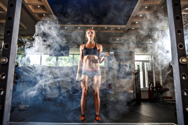 Sexy athlete blonde fitness woman in the gym workout with dumb on bicep. smoke - Zdjęcie, obraz