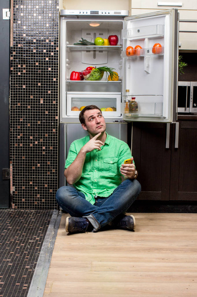 Young Man Sitting In Front Of Fridge - Foto, afbeelding