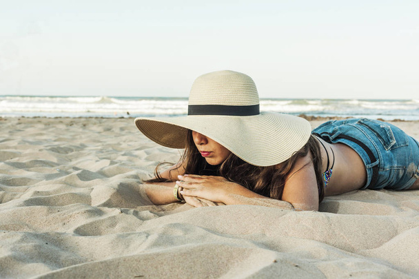 Girl laid on the beach wearing beach summer hat and short jeans. - Photo, Image