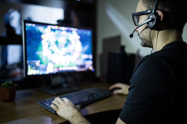  young gamer in headphones and glasses using computer for playing game at home - Photo, Image