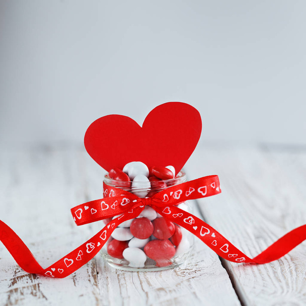 Colorful candy jar decorated with a red bow and red paper heart on white wooden background. Valentines day concept - Φωτογραφία, εικόνα