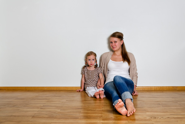 Mujer joven y niña sentada en el suelo de madera contra la pared blanca
 - Foto, imagen