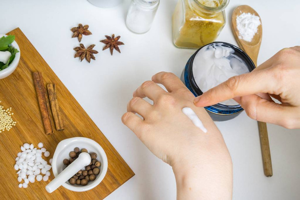 Young woman is applying homemade cream on her skin. A lot of ingredients for home made cosmetics in background. - Фото, зображення