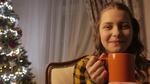 Teen girl smiling and drinking tea from a cup. Christmas tree and holidays mood. 4K UHD. - Video, Çekim