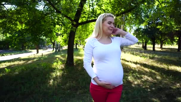Vrij vrouwelijke zwangere Blonde zachtjes strelen buik en brede glimlach, gegaap en rek, permanent in Green Park overdag in Open lucht. - Video