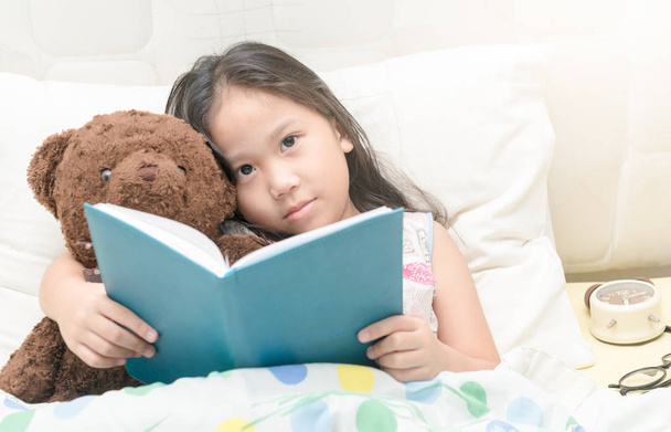 Söpö pieni aasialainen tyttö yöpaidassa lukeminen kirja nallekarhu b
 - Valokuva, kuva