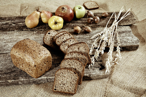 ψωμί σίκαλης με σπόρους σε ξύλινη σανίδα με μουσαμά και φρούτα γύρω - Φωτογραφία, εικόνα