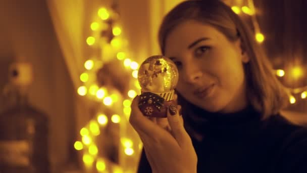 Portrait of a young woman with fairy lights in the house shaking the snow globe - Кадри, відео